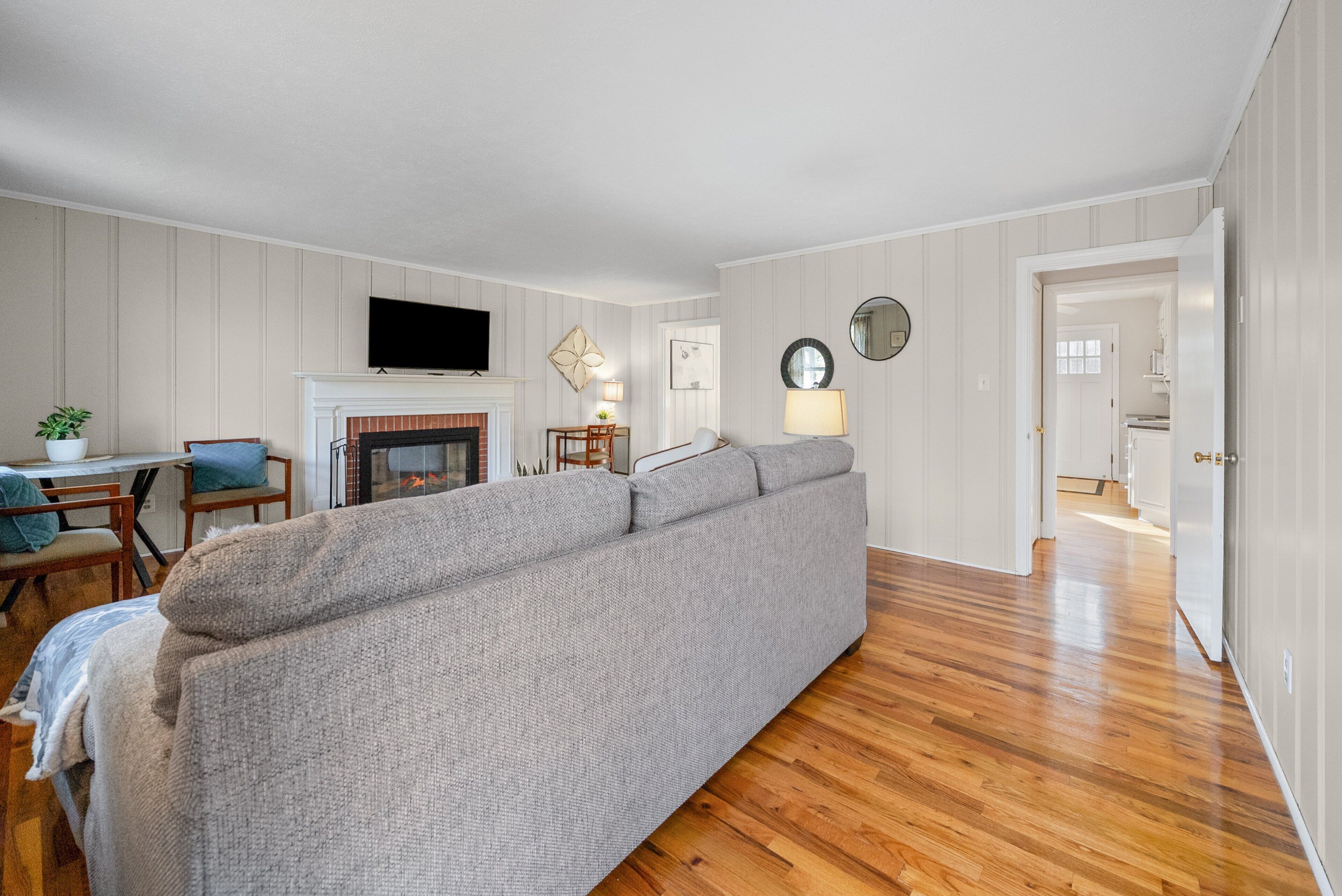 A living room with a grey sofa, wooden flooring, a fireplace, a TV mounted on the wall, and a dining area with a table and chairs.