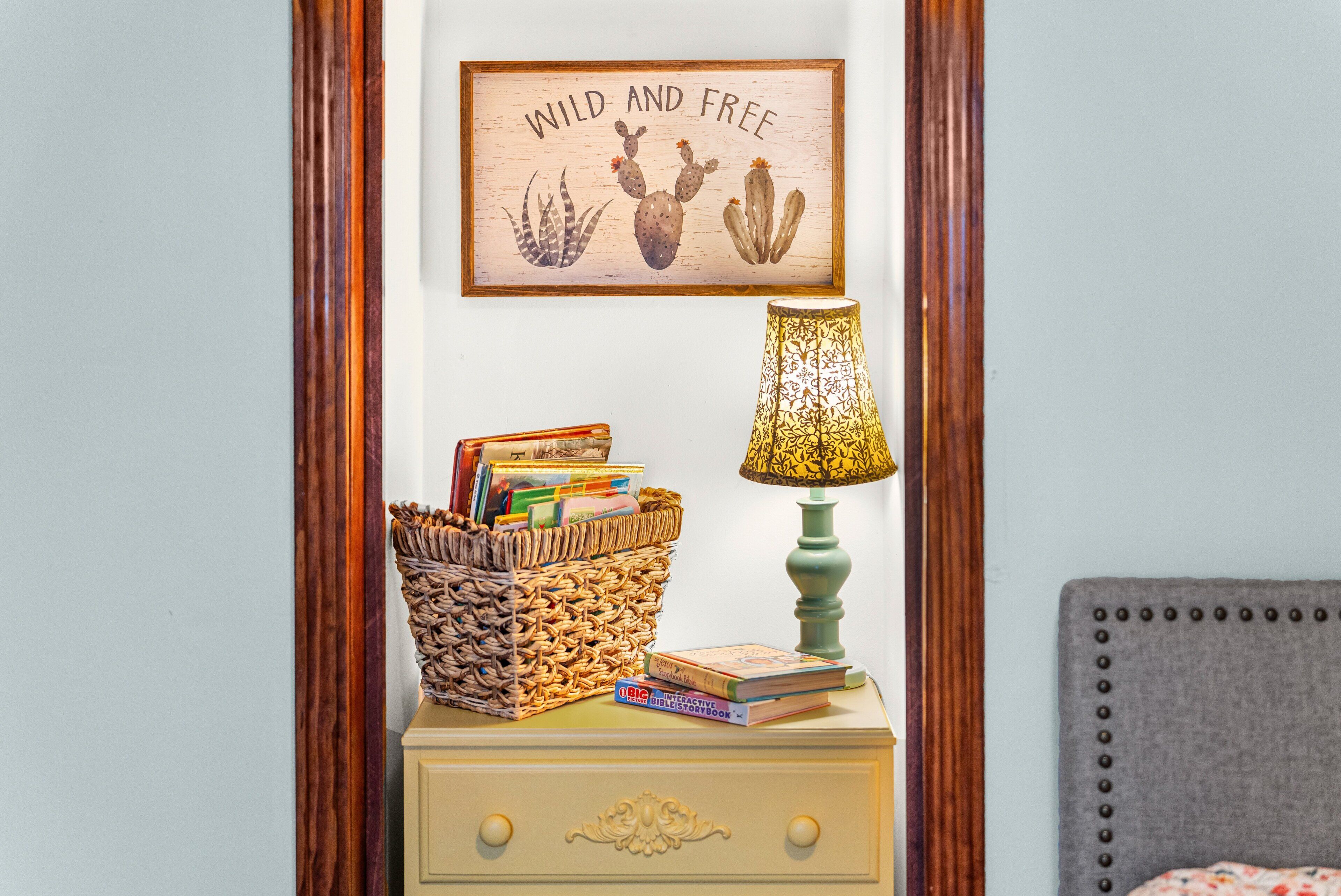 A bedside table with a woven basket, books, a lamp, and a framed picture on a wall.