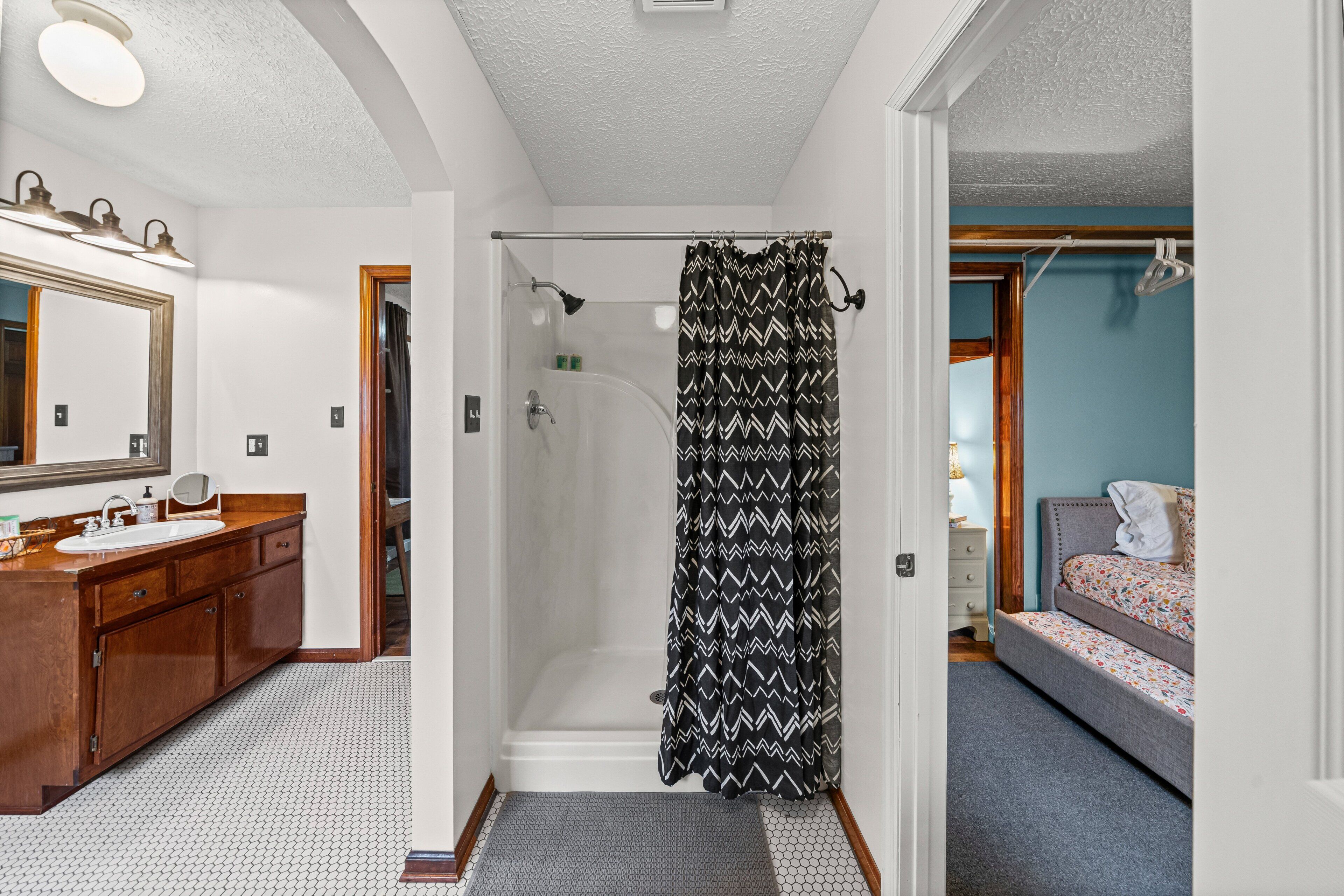 A bathroom with a shower, a sink, a mirror, and a door leading to a bedroom.