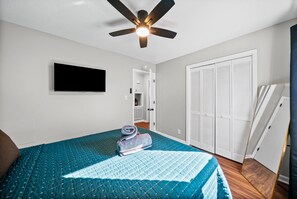 Une chambre à coucher comprenant un lit, un ventilateur de plafond, une télévision, un placard et une porte donnant sur une autre pièce.