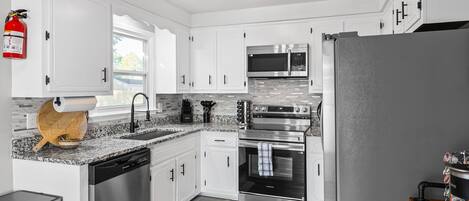 A modern kitchen with white cabinets, stainless steel appliances, a fire extinguisher, and a wooden cutting board.