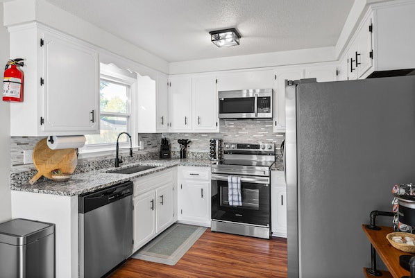 A modern kitchen with white cabinets, stainless steel appliances, a fire extinguisher, and a wooden cutting board.