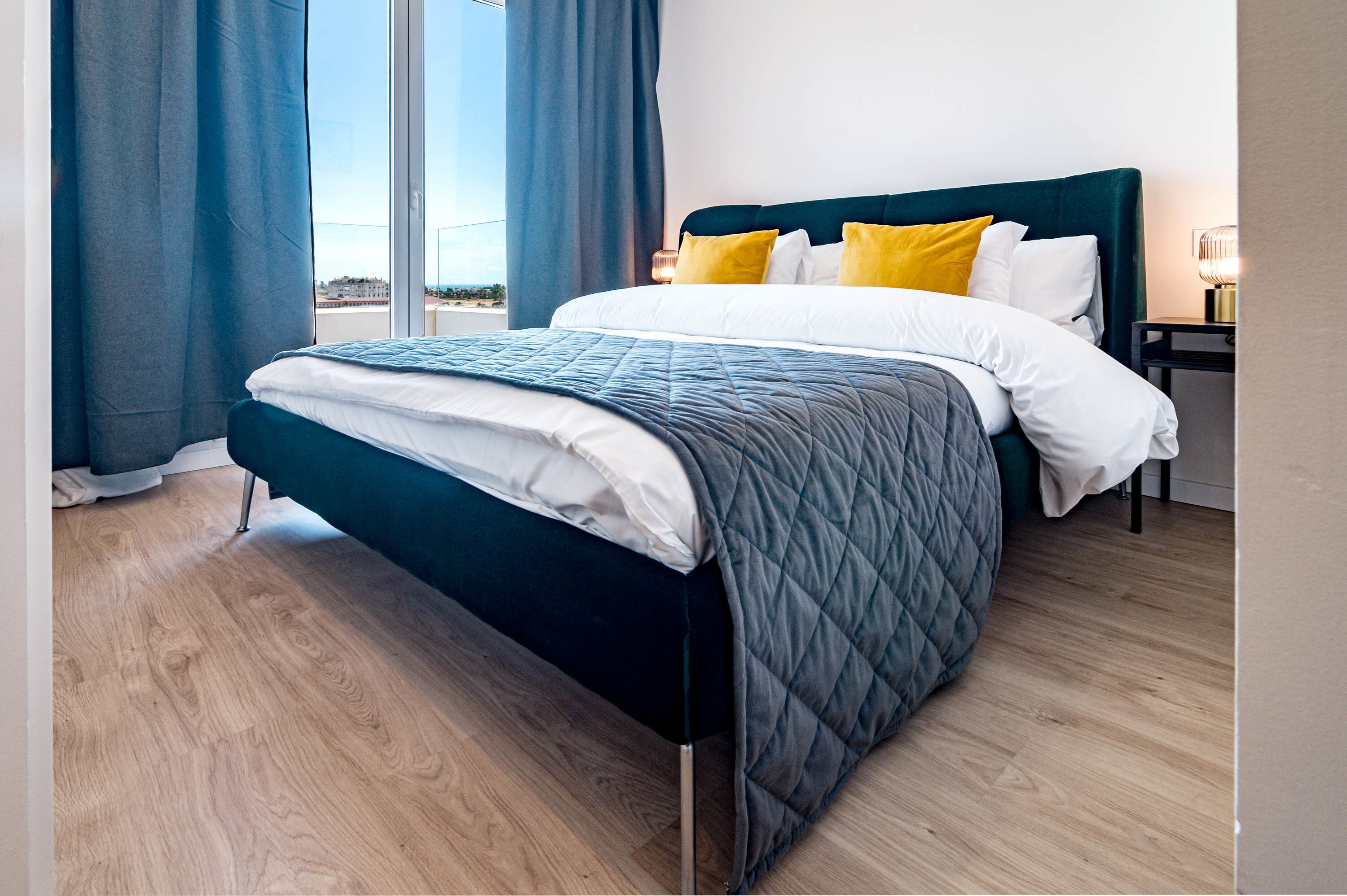 A modern hotel room with a large bed, a view through the window, and a nightstand with a lamp.