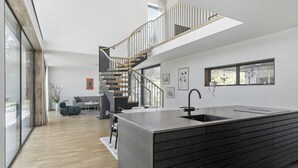 A modern kitchen with a central island, a staircase leading to an upper level, and a dining area with a sofa and table. - Airy house with large glass fronts on Adelsö (Adelsö)