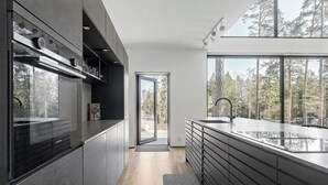 A modern kitchen with a large island, built-in appliances, and a view of a forest. - Airy house with large glass fronts on Adelsö (Adelsö)