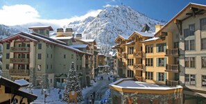 A snow-covered mountain town with multi-story buildings, a central pedestrian area, and a prominent snow-covered tree. - Alpenglow Loft | Palisades Adventure Basecamp (Olympic Valley)