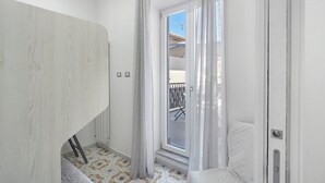 Una habitación con una puerta blanca, una ventana con cortinas, un piso estampado y una cama con cubrecama blanco.