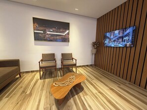 Une pi èce moderne avec un plancher en bois, deux fauteuils, un canapé, une table basse en bois ornée d’un collier de perles, un téléviseur fixé au mur et un grand tableau encadré.