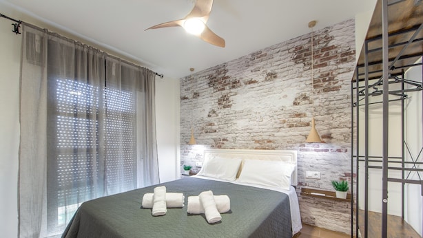 Un dormitorio con cama, ventilador de techo, pared de ladrillo y ventana con cortinas.