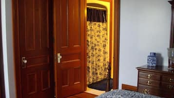 Une chambre avec une porte en bois, un lit recouvert d’une couvre-lit à motifs floraux, une commode avec des tiroirs et une petite table sur laquelle est posé un vase.