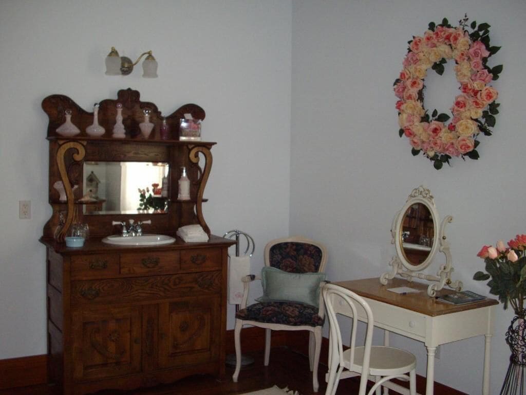 A room with a wooden dresser, a chair, a table with a mirror, and a floral wreath on the wall.