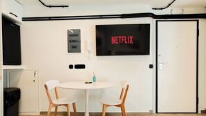 Ein modernes Zimmer mit einem runden Tisch, zwei Stühlen, einem Fernsehbildschirm, der 'NETFLIX' anzeigt, einer kleinen Ablage mit einer Kerze und einer Tür mit Griff.