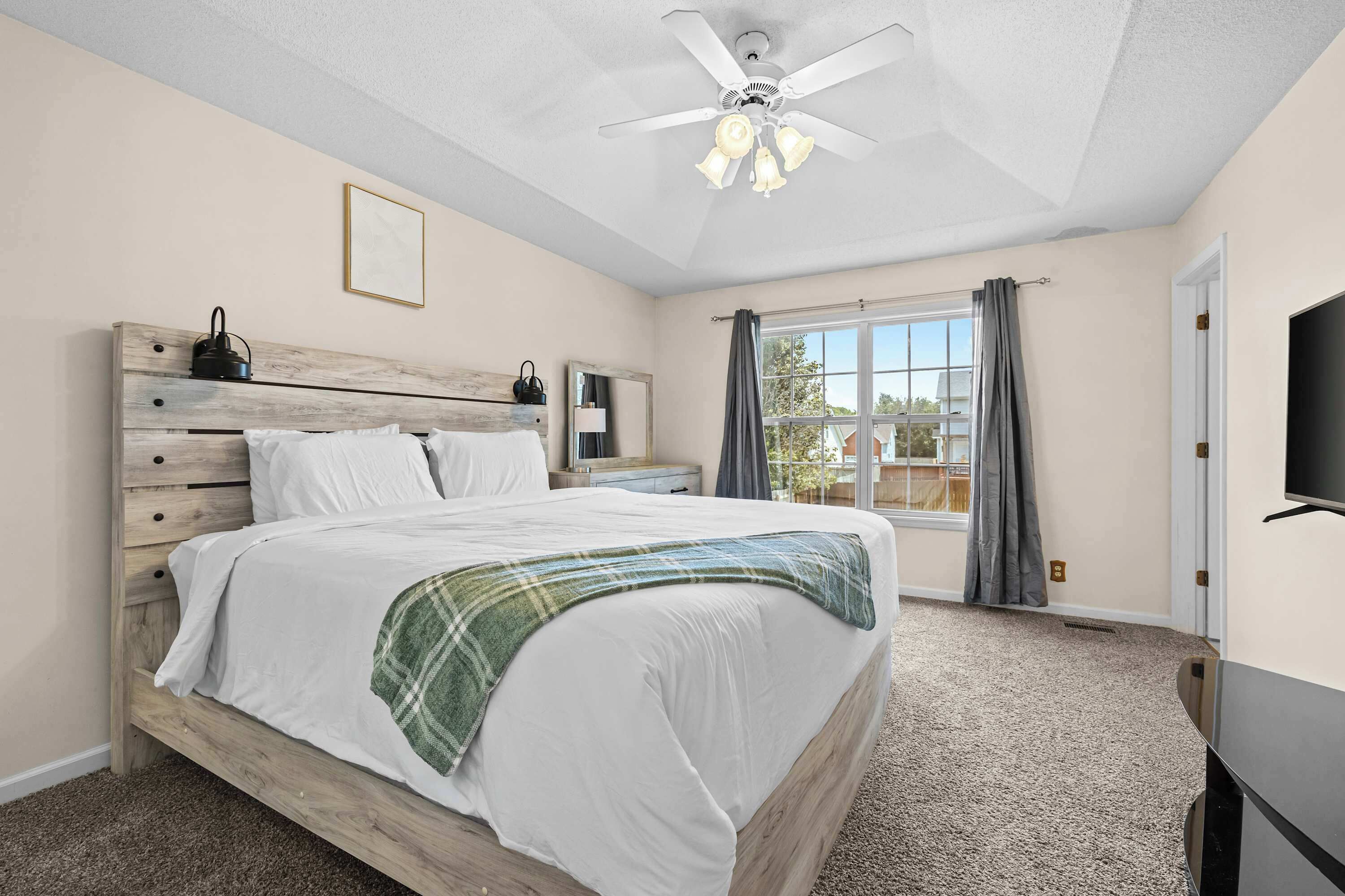 Une chambre à coucher avec un lit, un ventilateur de plafond, une fenêtre avec des rideaux et un téléviseur fixé au mur.