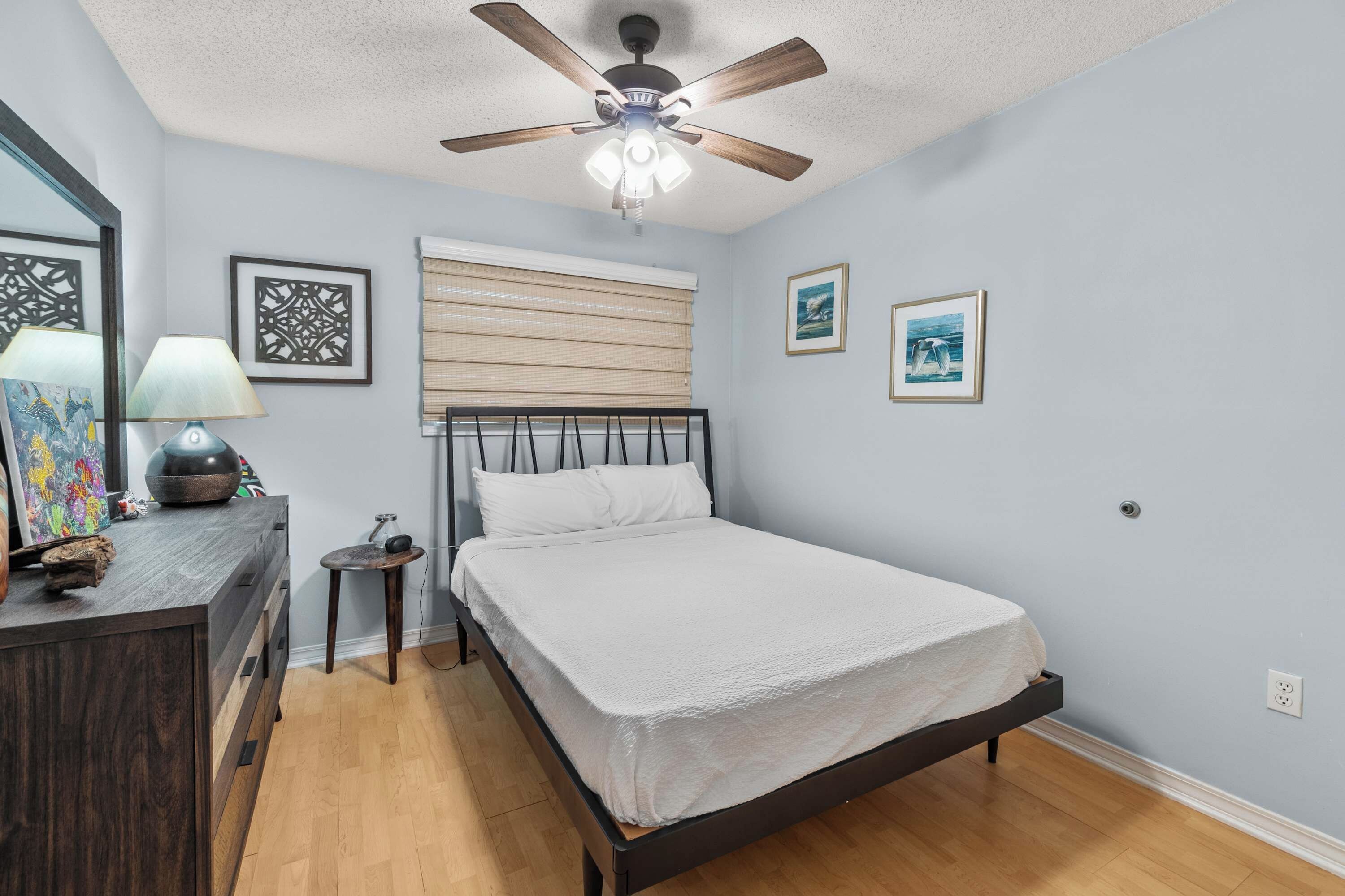 Un dormitorio con cama, ventilador de techo, mesita de noche, lámpara y decoración en la pared.