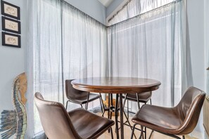 Quarto com uma mesa redonda e quatro cadeiras, uma janela com cortinas leves e uma parede com obras de arte emolduradas.