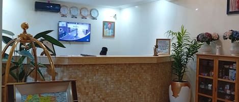 A reception area with a check-in counter, a wall-mounted TV, clocks, a framed map, and a potted plant.