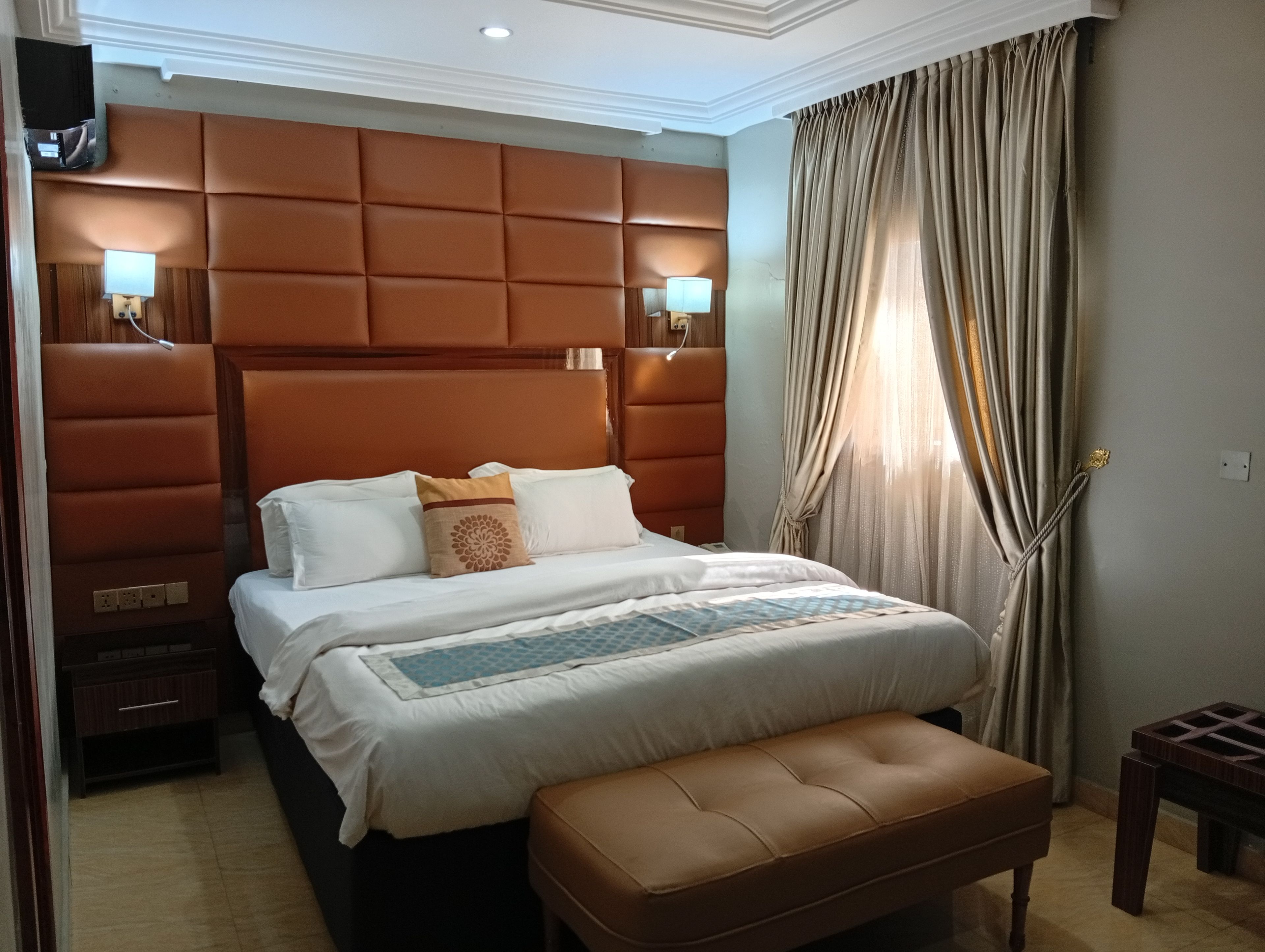 Une chambre d’hôtel avec un grand lit, une tête de lit, des lampes de chevet, un banc et une fenêtre avec des rideaux.
