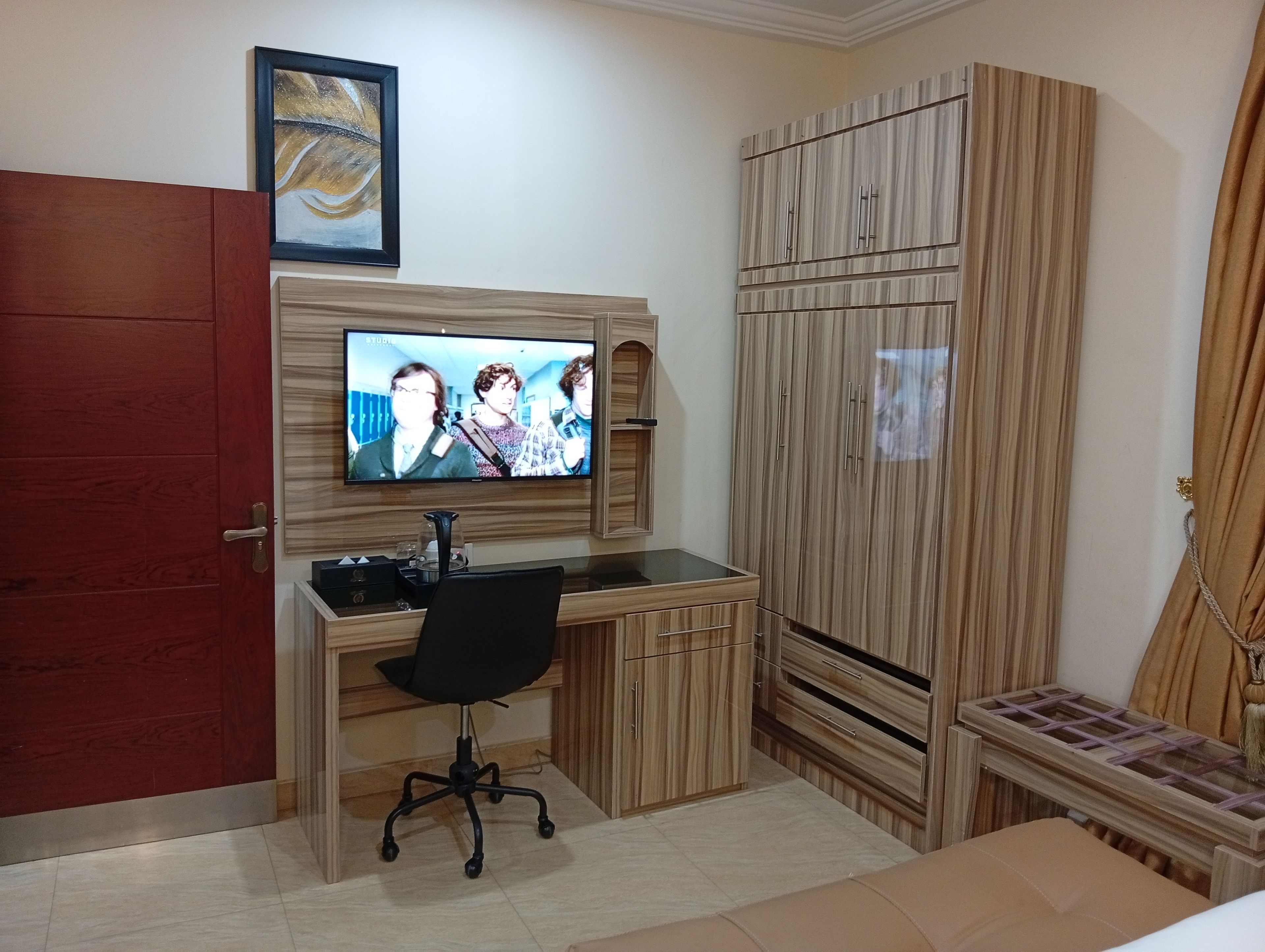 A bedroom with a TV mounted on the wall, a wooden wardrobe, a desk with a chair, and a bed.