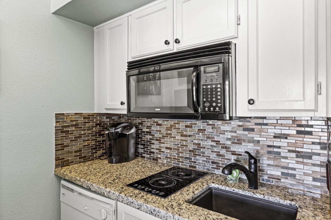 A kitchen with white cabinets, a black microwave, a coffee maker, a stovetop, and a tiled backsplash.