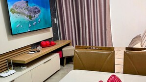 Un salon moderne doté d’un téléviseur à écran plat, d’un meuble de rangement en bois, d’une table à manger recouverte d’une nappe blanche et d’une grande fenêtre agrémentée de rideaux.