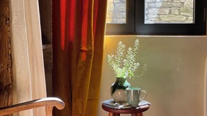 A room with a window, a chair, a small stool, a vase with flowers, and a stone wall.
