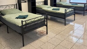 Een slaapzaal met drie metalen stapelbedden, elk voorzien van een groene matras en bijpassende kussens. Boven elk bed hangen wandplanken, en aan de verre wand is een raam met jaloezieën.