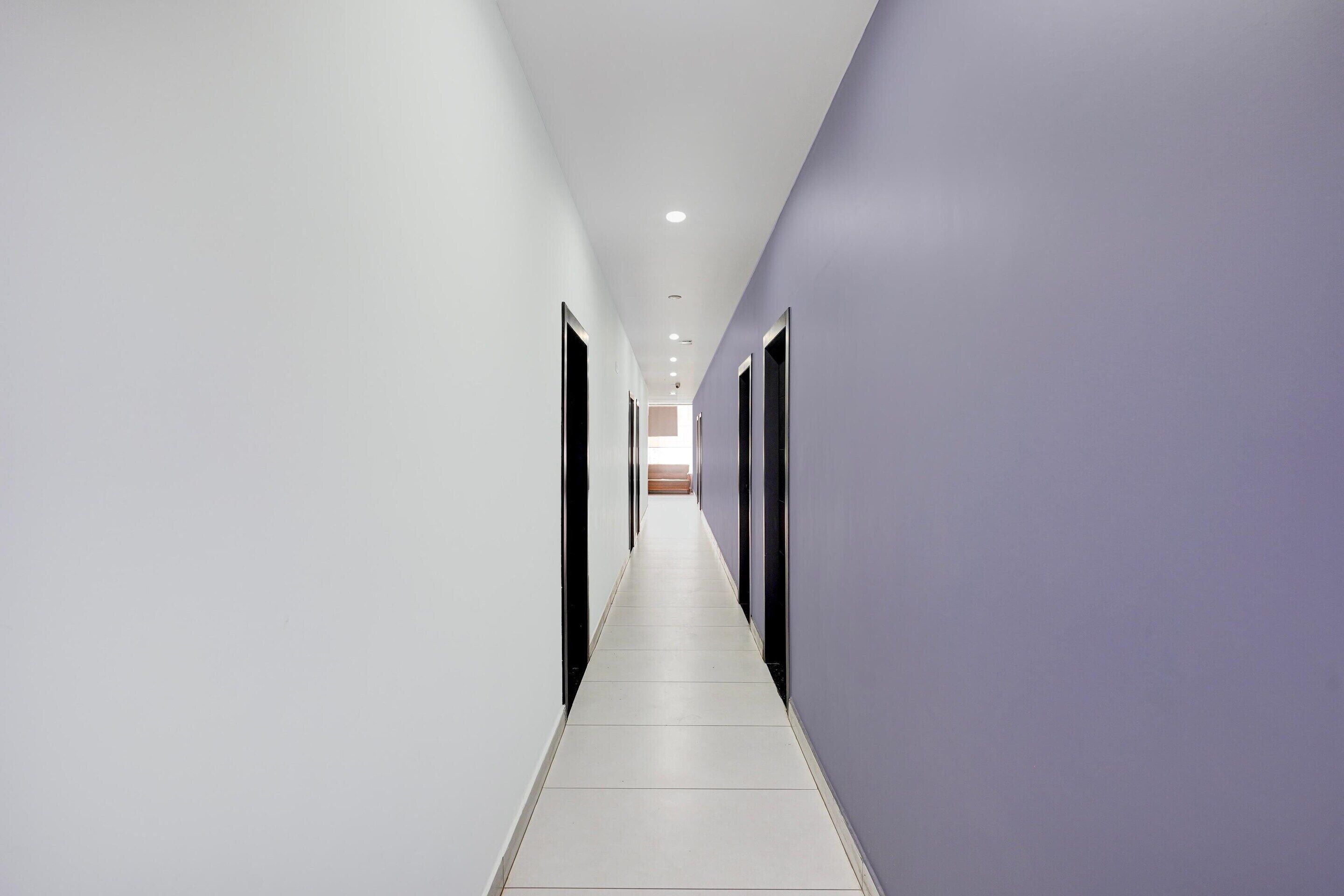 A long, narrow hallway with white walls and a tiled floor, leading to a room with a large window.