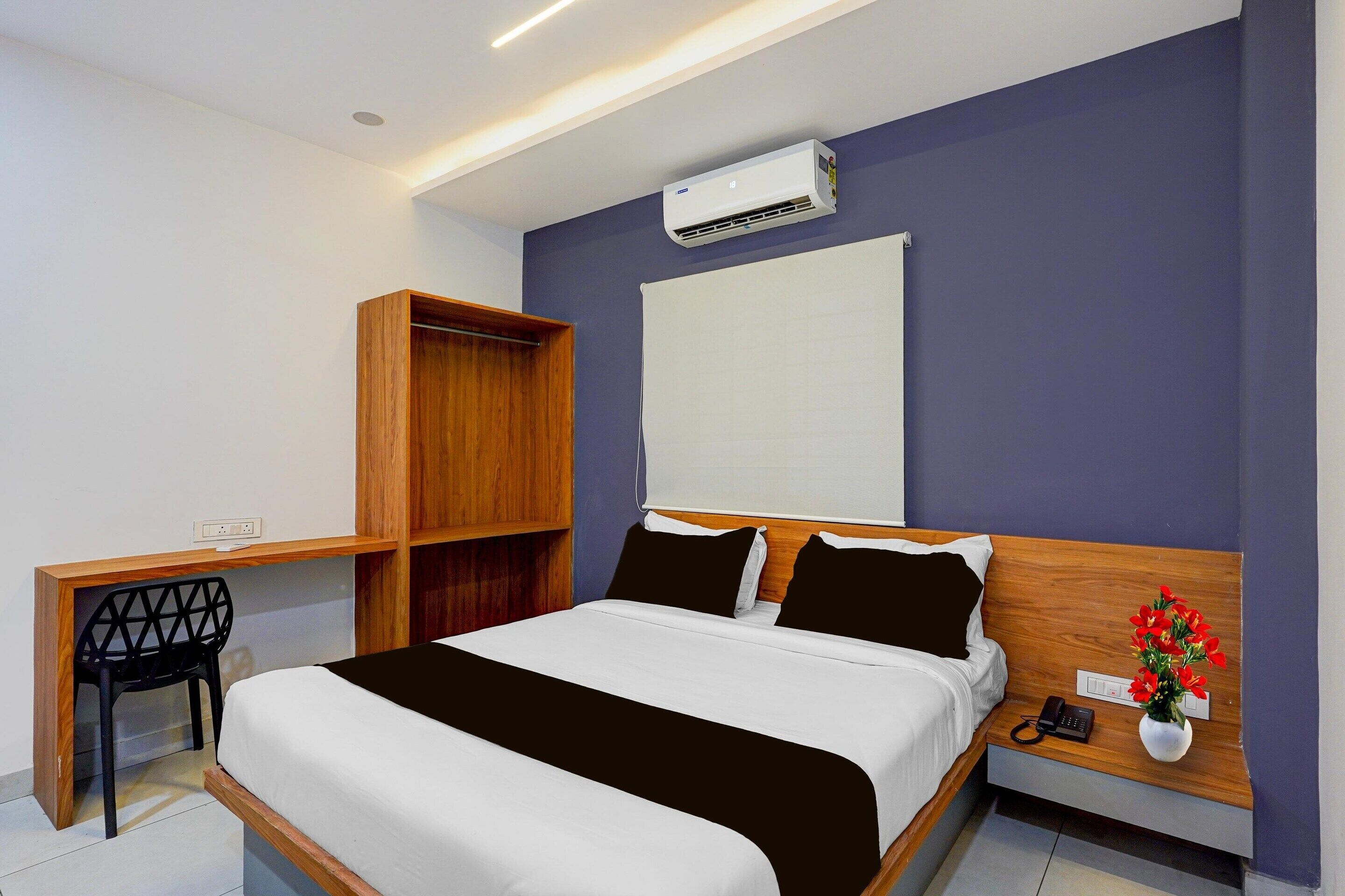 A hotel room with a bed, a desk, a wardrobe, an air conditioning unit, a bedside table with a phone and flowers, and a window with blinds.