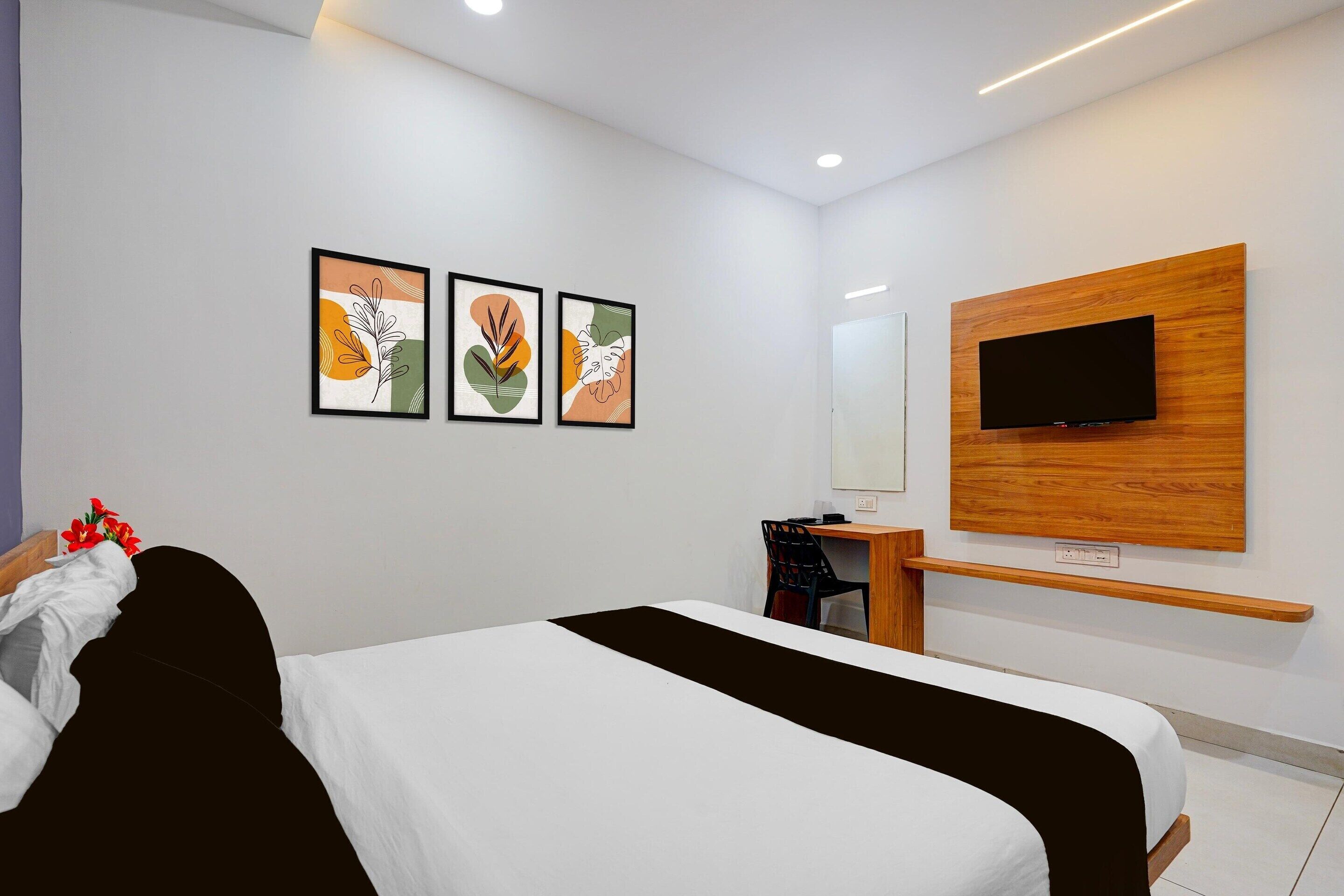 A hotel room with a bed, a desk, a chair, a TV mounted on a wooden panel, and three framed artworks on the wall.