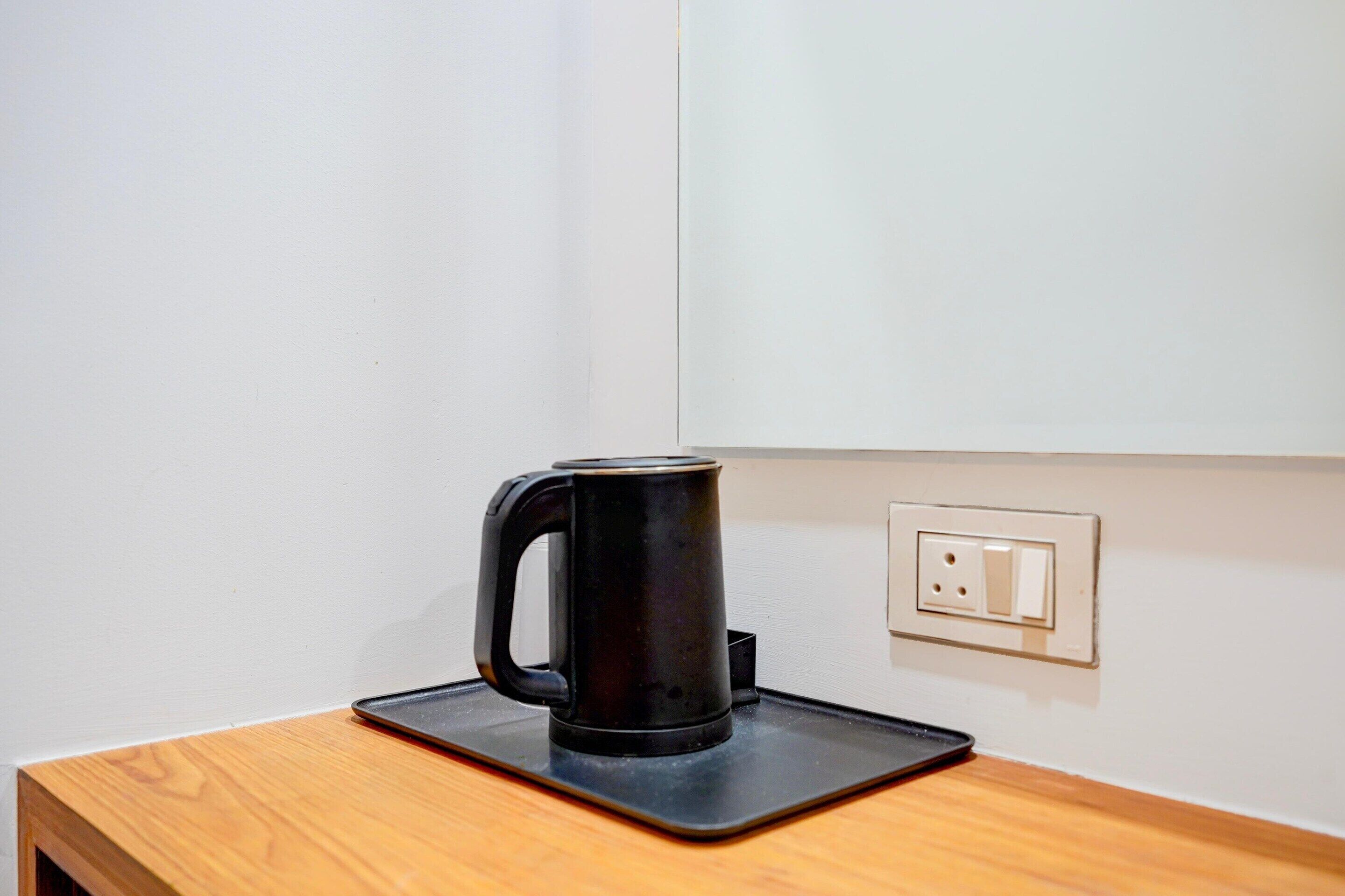 Una cafetera negra sobre un posavasos negro en una mesa de madera, frente a una pared blanca con un enchufe.
