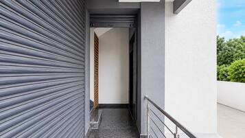 Un estrecho pasillo exterior con barandilla metálica, un persiana enrollable cerrada y una pared blanca.