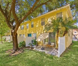 Un bâtiment jaune doté d’un espace extérieur aménagé, couvert et entouré d’arbres et d’herbe.
