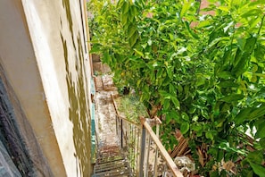 Un estrecho pasillo exterior con barandilla metálica, rodeado de abundante follaje verde y un camino de piedra.
