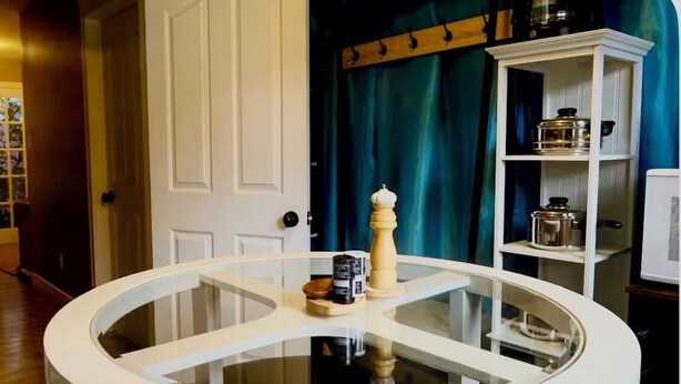 Ein Zimmer mit einem Glastisch, einer weißen Tür, einem blauen Vorhang und einem weißen Regal mit Küchenutensilien.