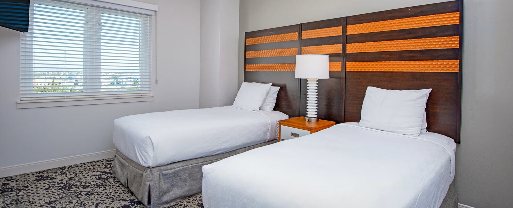 A hotel room with two beds, a window with blinds, a nightstand with a lamp, and a patterned carpet.