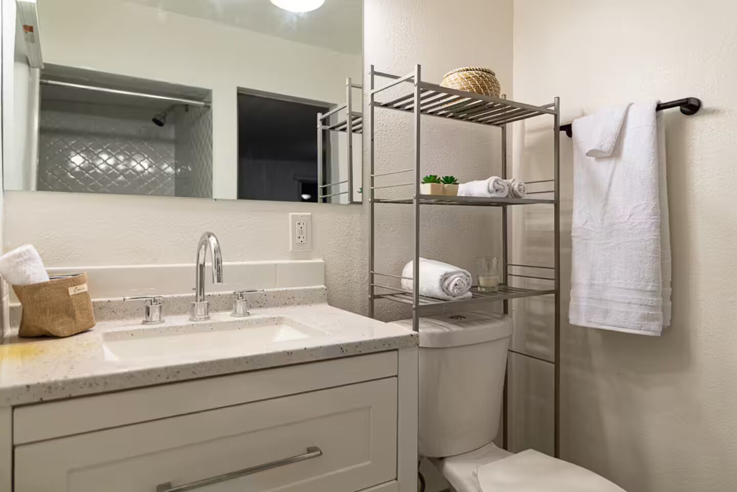 Un baño con lavamanos blanco, espejo, toallero y estante metálico.