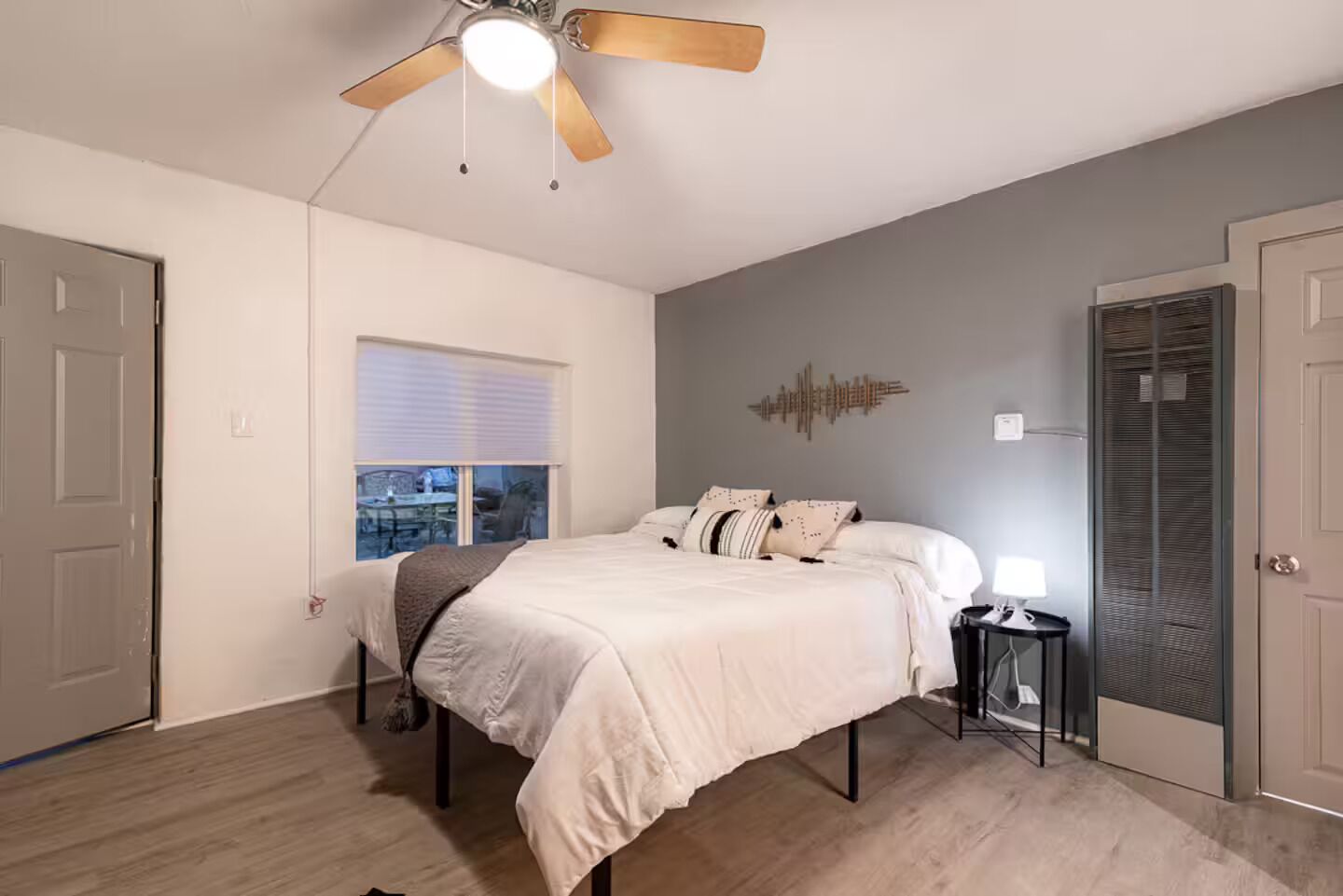 Un dormitorio con cama, ventilador de techo, ventana, un cuadro en la pared y una puerta.