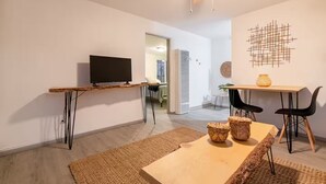 Een moderne woonruimte met een televisie op een houten standaard, een eettafel met stoelen, een plafondventilator en een kunstwerk aan de muur.