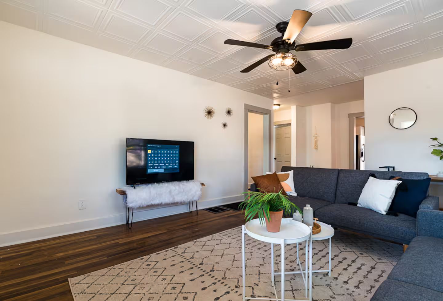 Un soggiorno moderno con TV a schermo piatto, ventilatore a soffitto, divano grigio, tavolino bianco e tappeto decorato.