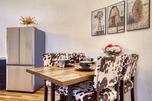 Un coin repas avec une table en bois, deux chaises recouvertes d’un tissu à motifs, un réfrigérateur et trois tableaux encadrés accrochés au mur.