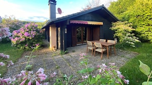 A small house with a chimney, a covered patio with a table and chairs, and a garden with flowering plants.