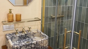 A bathroom with a marble sink, a mirror, a shelf with decorative items, and a glass-enclosed shower.