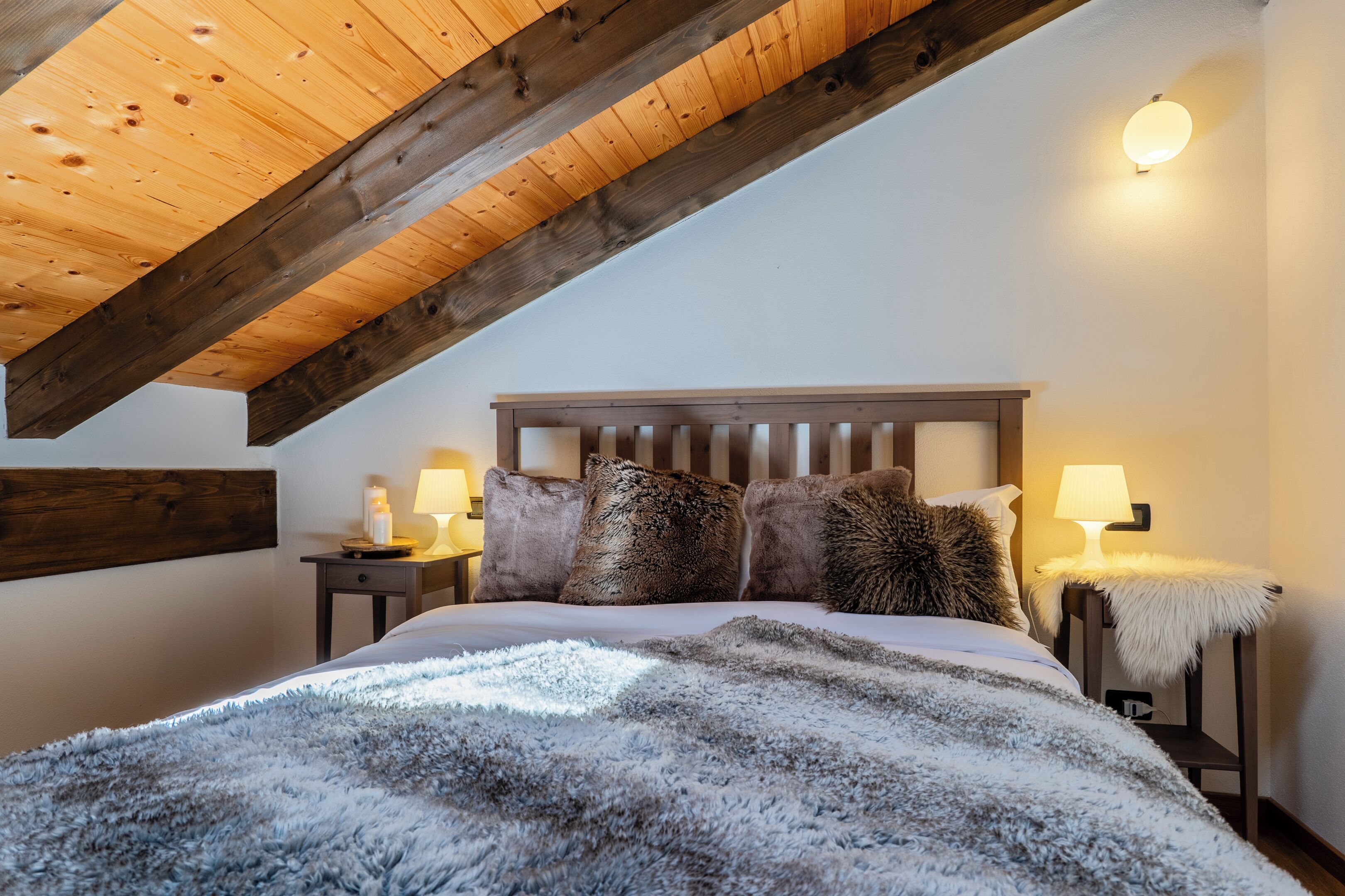 A bedroom with a sloped ceiling, wooden headboard, two bedside tables with lamps, and a fluffy blanket.