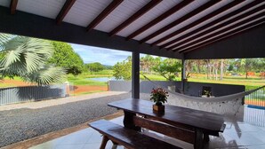 A covered outdoor area with a wooden table and bench, a hammock, and a view of a green landscape with trees and a body of water.