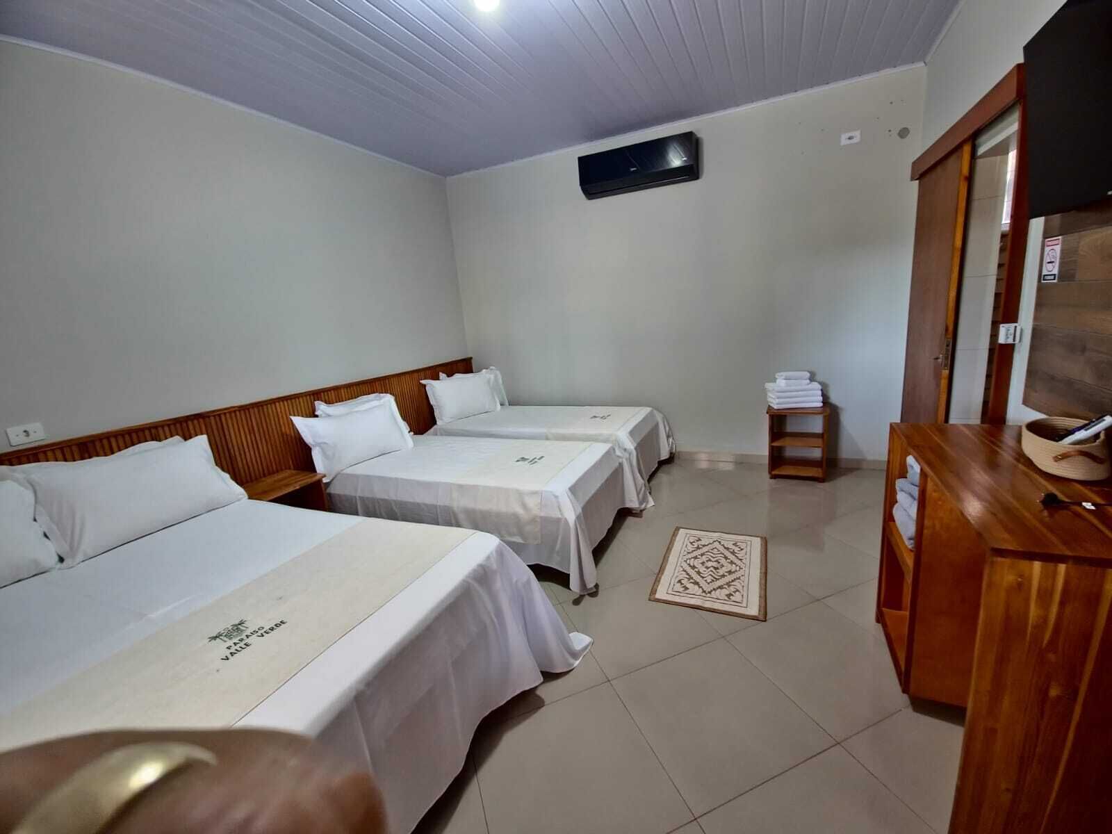 Quarto de hotel com três camas, cabeceira de madeira, uma mesinha com um cesto e ar condicionado fixado na parede.