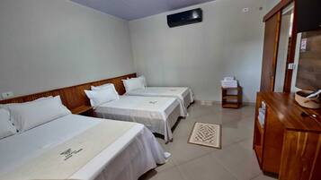 Quarto de hotel com três camas, cabeceira de madeira, uma mesinha com um cesto e ar condicionado fixado na parede.