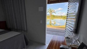 A room with a bed, a wooden desk, a remote control, and a view of a lake and palm trees through an open door.