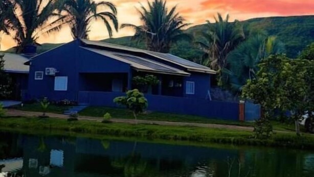 A house with a blue exterior, a pond in the foreground, and palm trees in the background.
