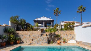 A pool area with a stone wall, palm trees, and a building with a tiled roof. - CH Villa Aström La Marquesa Golf (Ciudad Quesada)