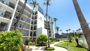 A multi-story hotel with a white facade, balconies, and a landscaped garden with palm trees and shrubs. - Beachfront 2BR, 5th Floor Views, Wakable SPI (South Padre Island)
